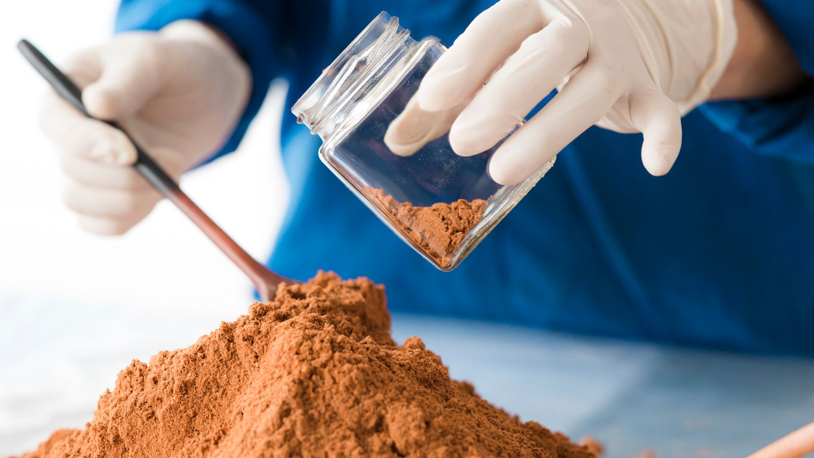 A scientist in blue gloves and attire examines a fine powder sample with a small wooden spoon on a lab bench.