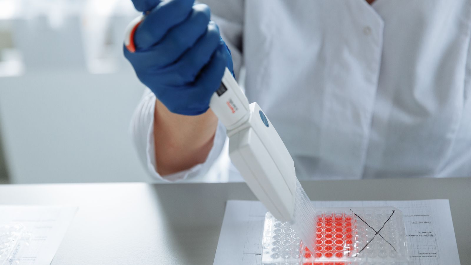 A gloved hand holds a pipette above a lab bench, drawing liquid from a small vial under focused lighting.