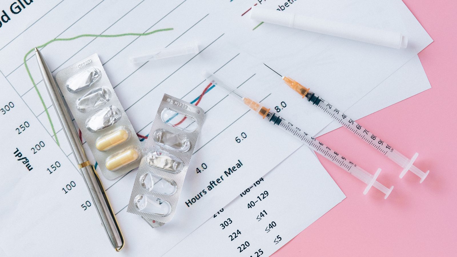 A flat lay of medicine vials, syringes, and a blood glucose chart arranged on a pink surface.